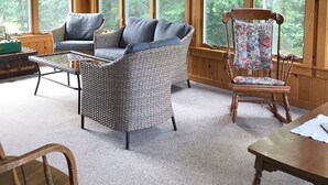 A cozy cabin interior with a wicker sofa, a rocking chair, and a wooden table. Large windows offer views of the surrounding forest.