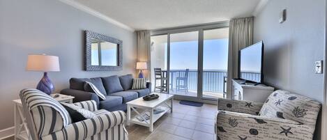 A modern hotel room with a balcony, a sofa, a striped armchair, a television, and a large mirror.