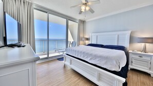 A bedroom with a large bed, a ceiling fan, a nightstand, a TV, and a view of the ocean through a sliding glass door.