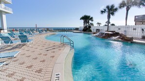 A pool area with lounge chairs, a pool with a waterfall feature, and a white fence with palm trees.