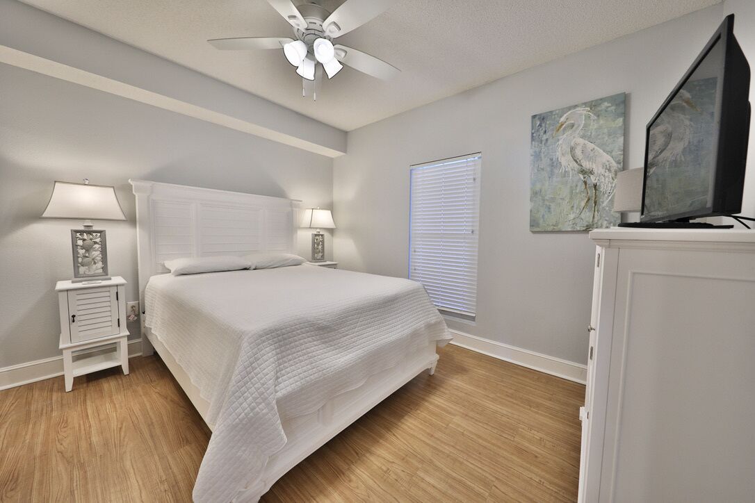 Une chambre à coucher avec un lit, des lampes de chevet, un ventilateur de plafond, une fenêtre avec des stores et un tableau au mur.