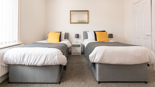 Habitación doble con dos camas, cada una con base gris y ropa de cama blanca, una mesita de noche con lámpara y un espejo en la pared.
