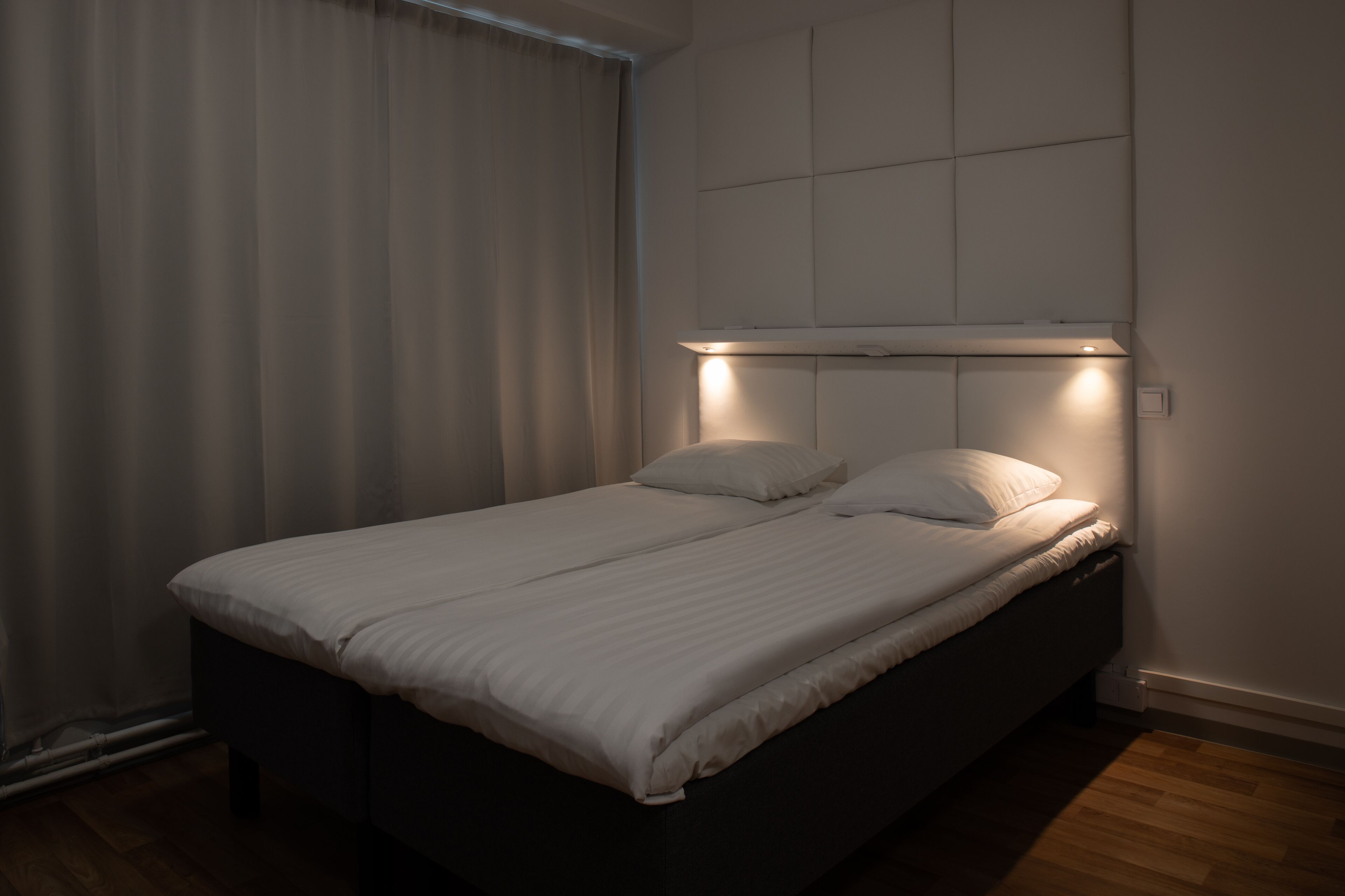 A neatly made bed in a room with a headboard, a window with curtains, and a wooden floor.