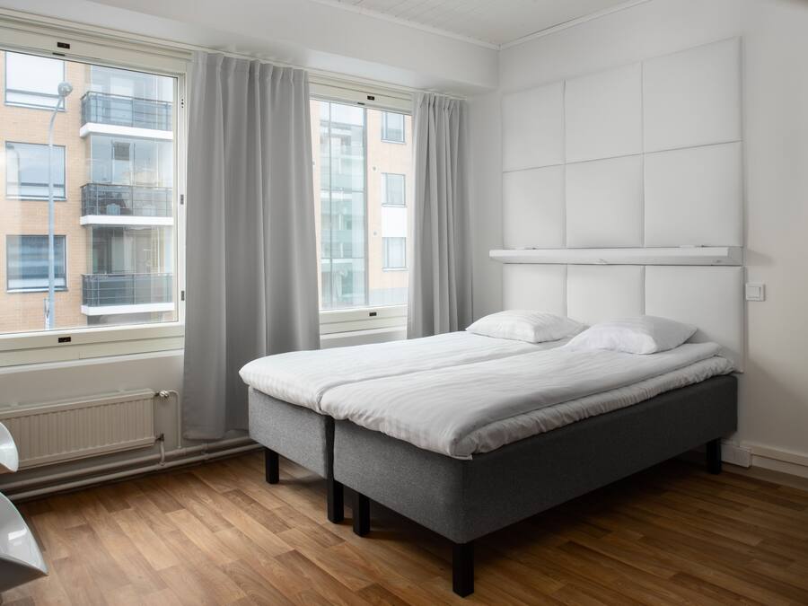 Quarto moderno com cama cinza, cabeceira branca e vista de um edifício através de janelas amplas.