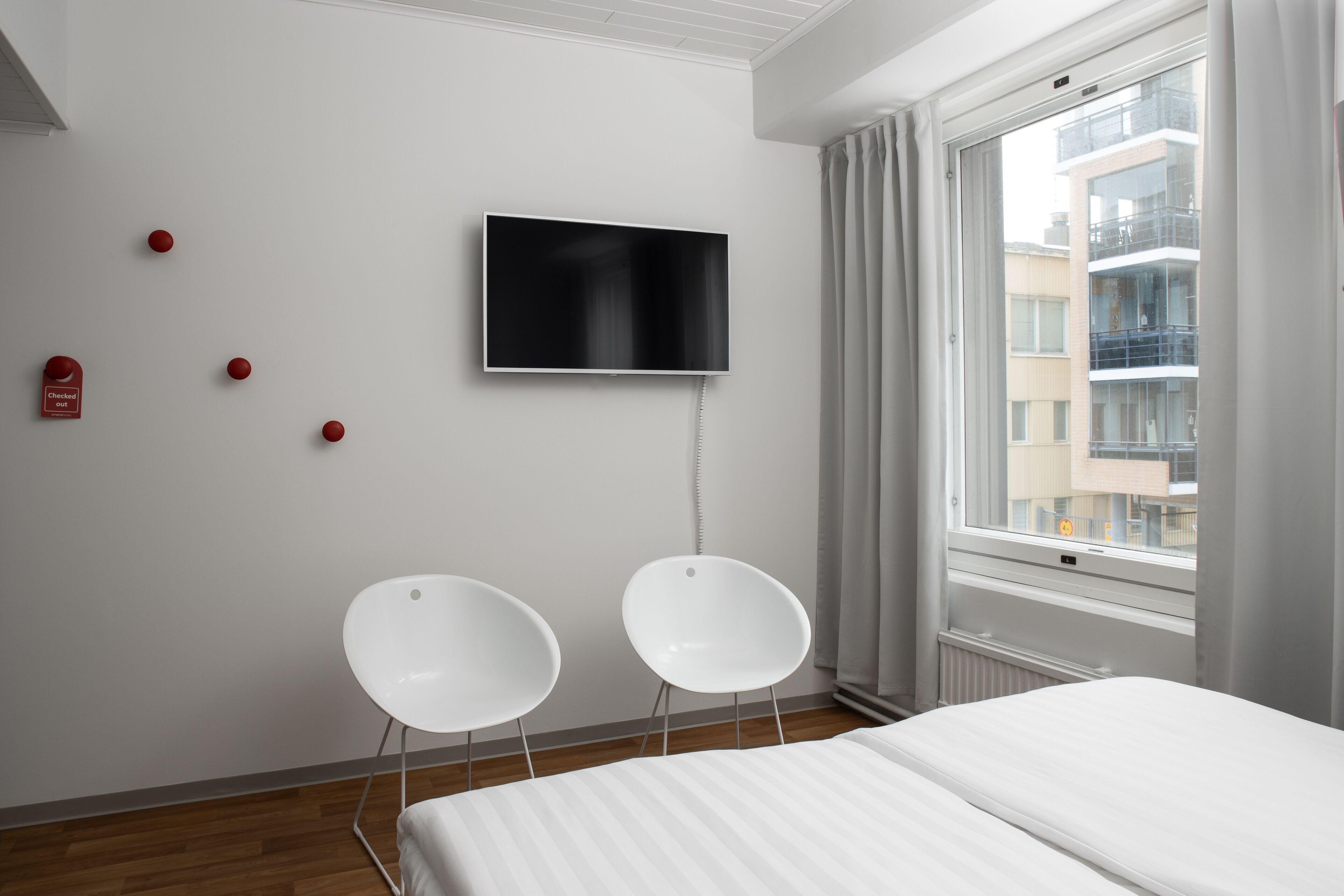 Quarto de hotel moderno com uma cama, duas cadeiras brancas, uma televisão de tela plana na parede e uma janela com vista para edifícios.