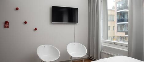 Quarto de hotel moderno com uma cama, duas cadeiras brancas, uma televisão de tela plana na parede e uma janela com vista para edifícios.