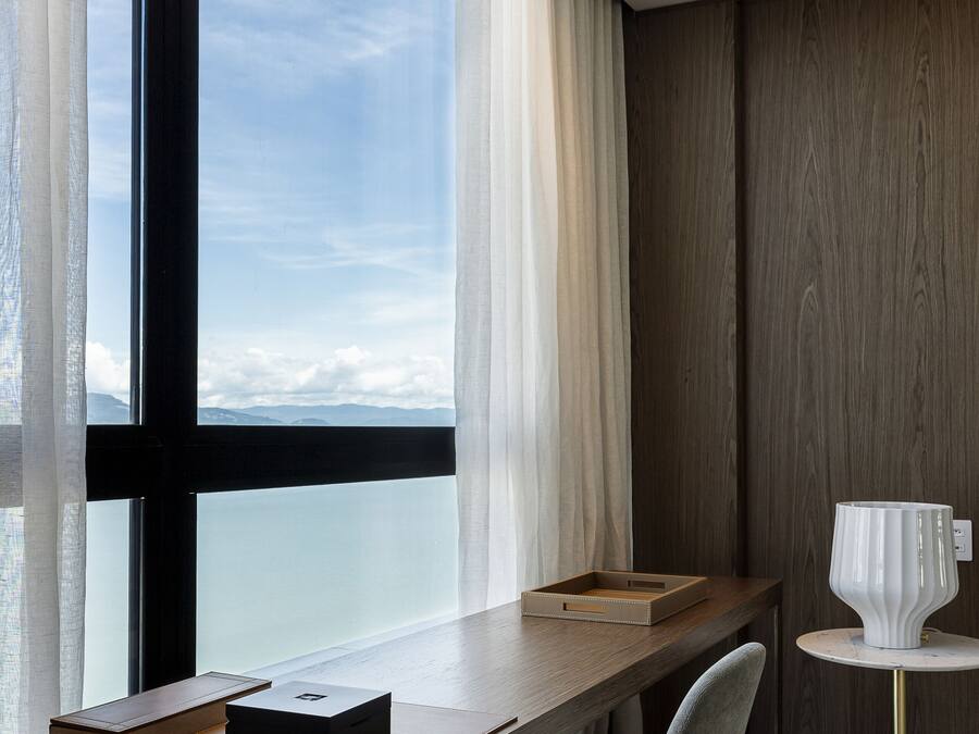 A modern hotel room with a large window, a wooden desk, a chair, and a small table with a lamp.