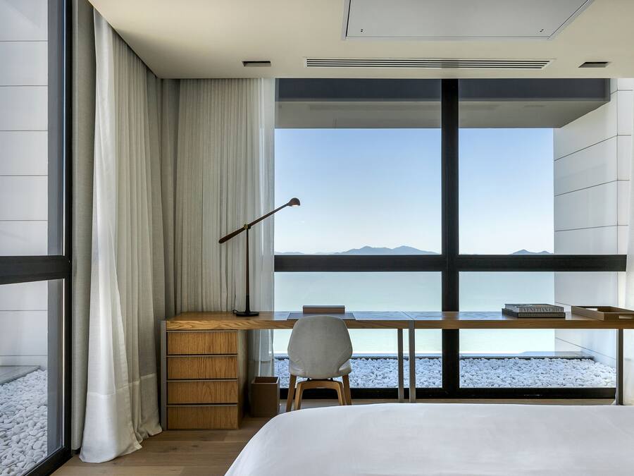 A modern hotel room with a large window, a wooden desk, a chair, a bed, and a nightstand.