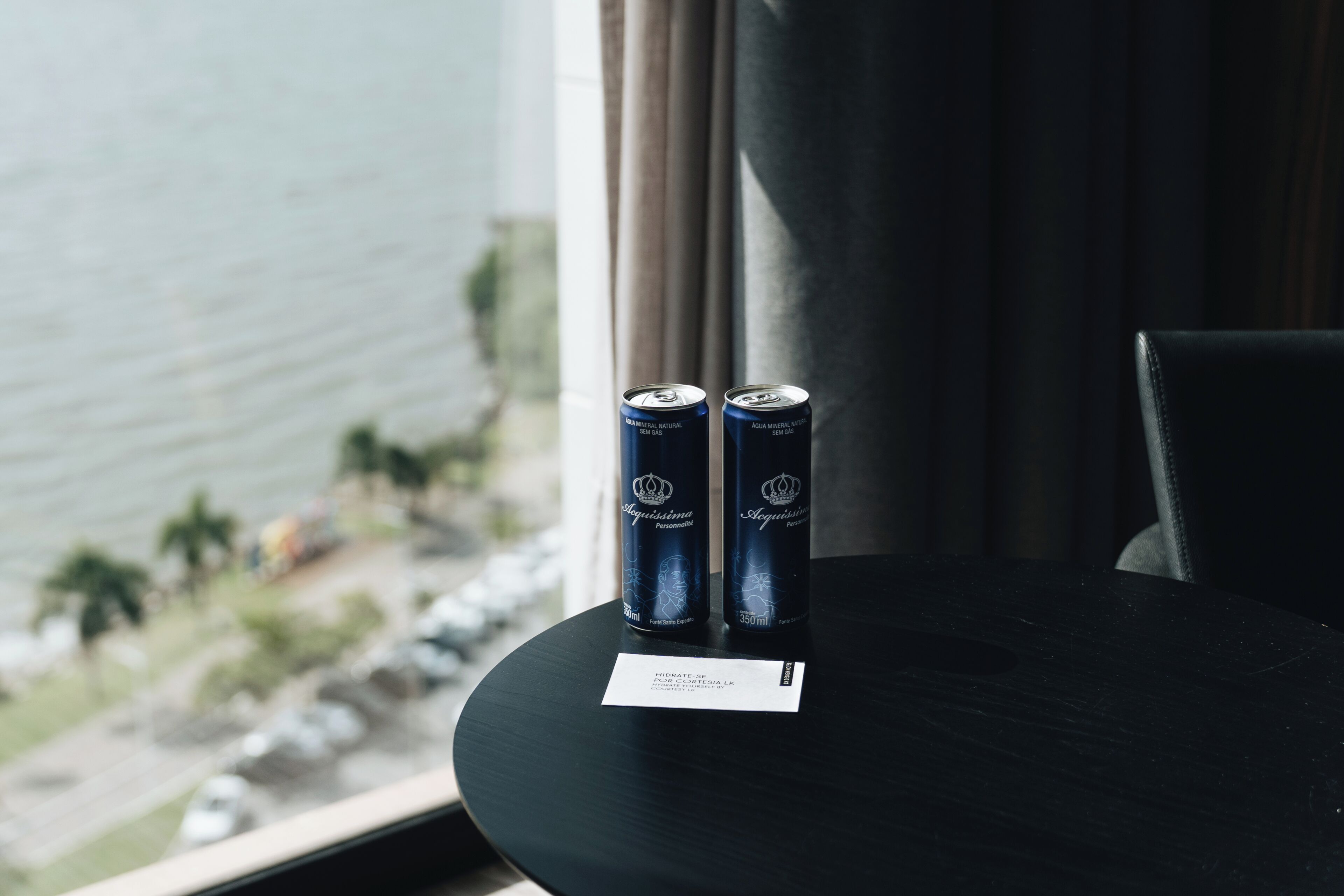Two cans of water on a table with a card, near a window overlooking a beach.