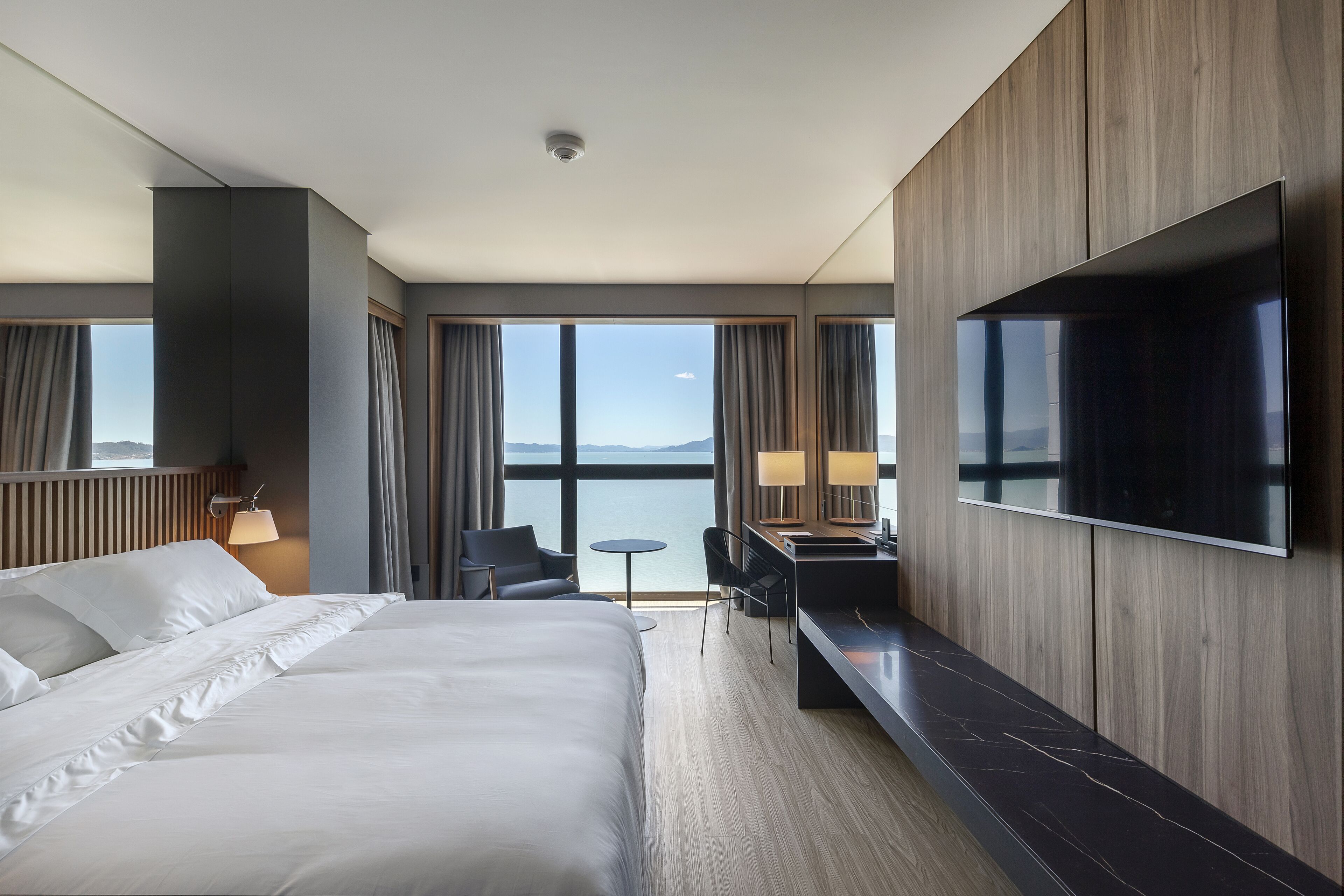 A modern hotel room with a large bed, a desk with a flat-screen TV, and a view of the sea.