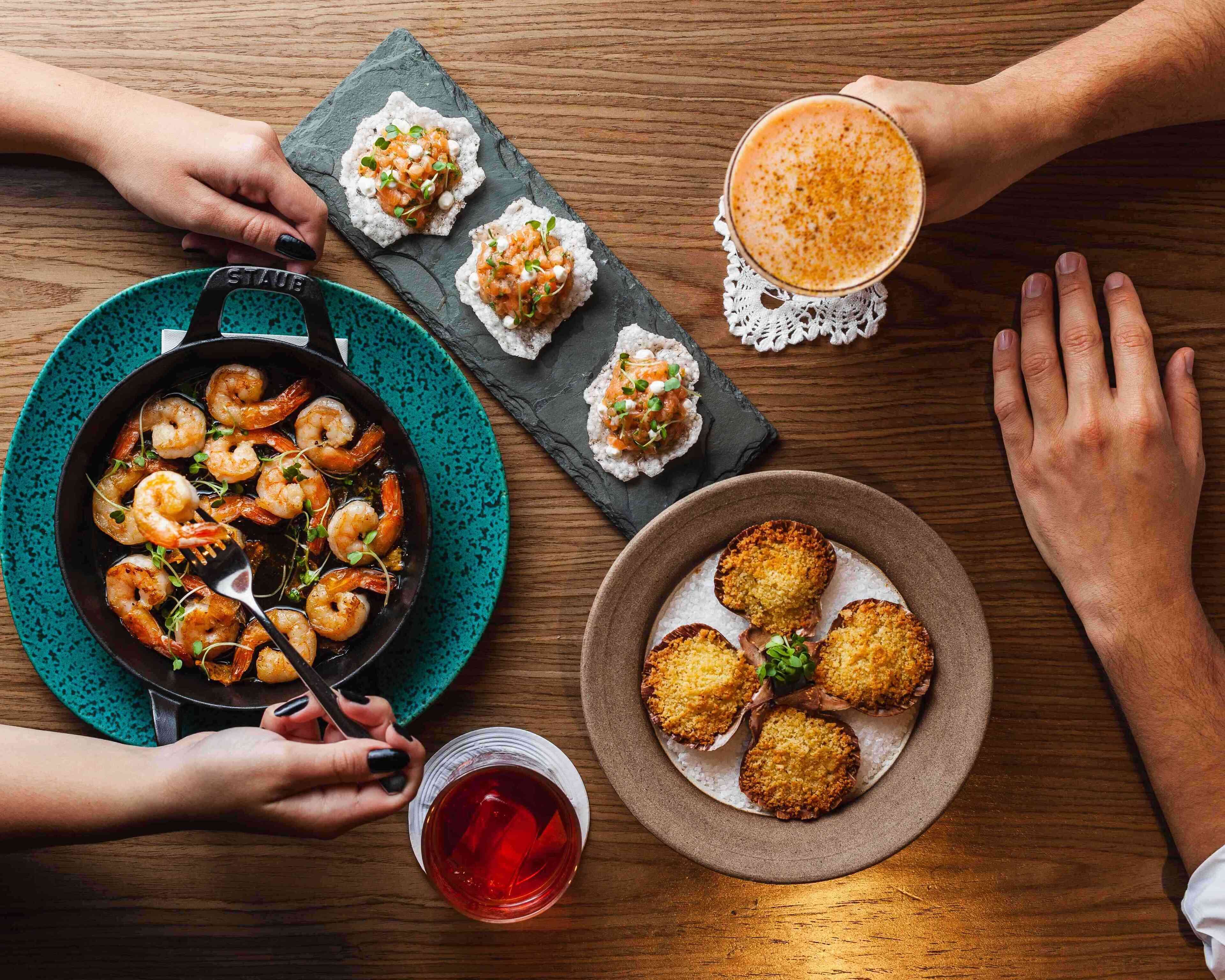 A dining table with a pan of shrimp, a plate of breaded items, a glass of coffee, and a cup of tea.