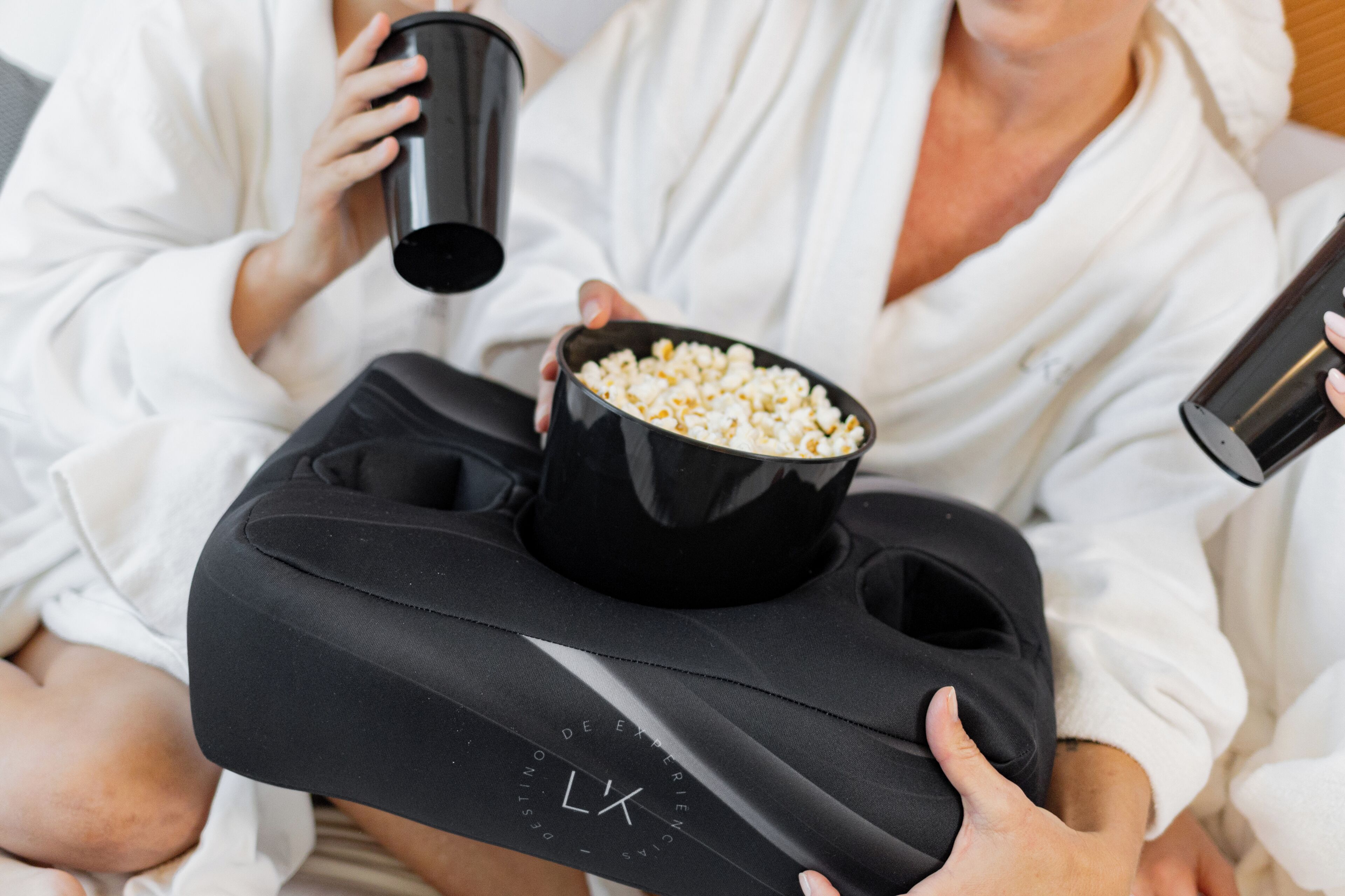 Una persona che tiene un vassoio nero con un sacchetto di popcorn e una tazza nera, vestita con una veste bianca.
