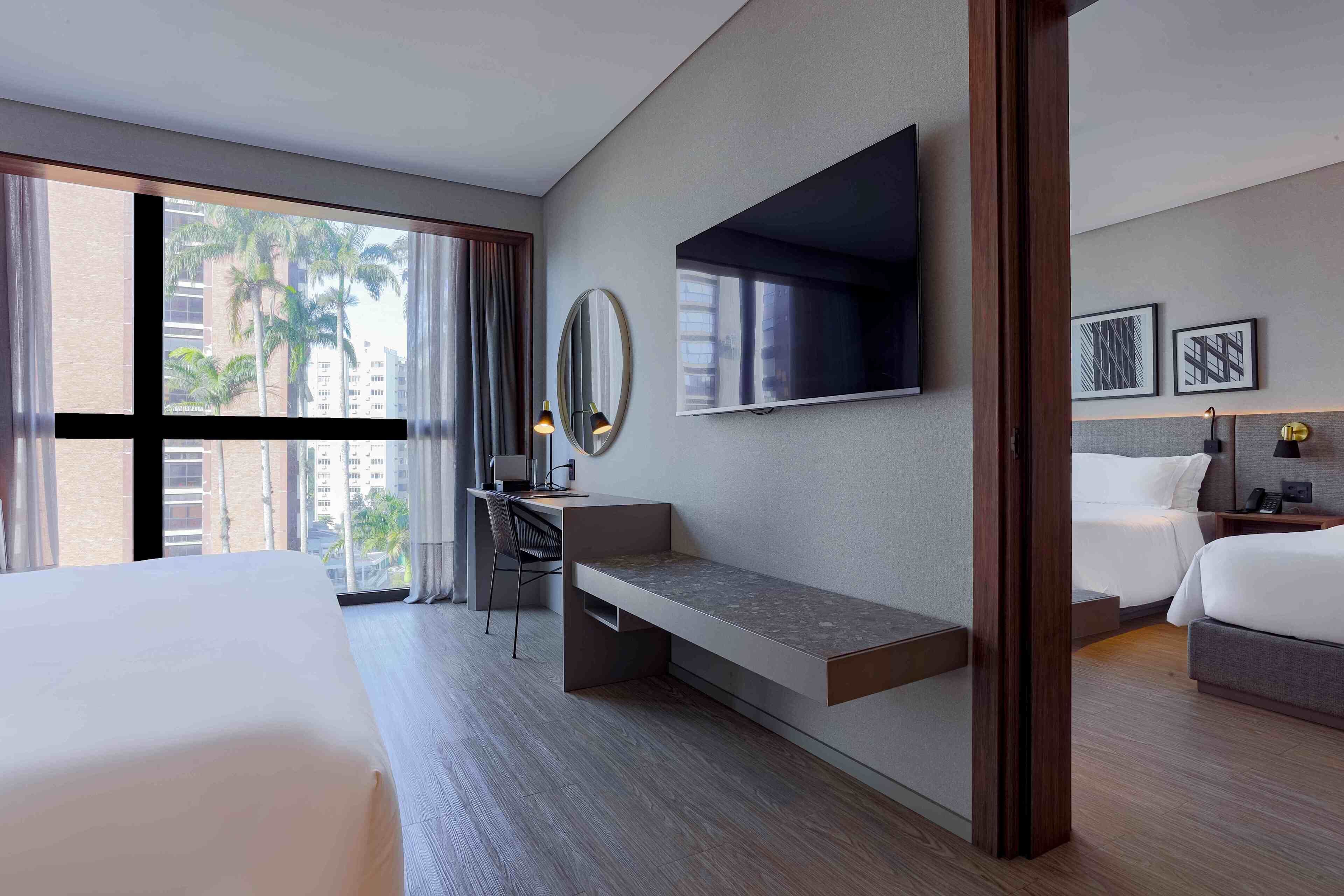 A modern hotel room with a large bed, a desk with a chair, a television, and a view of palm trees through the window.