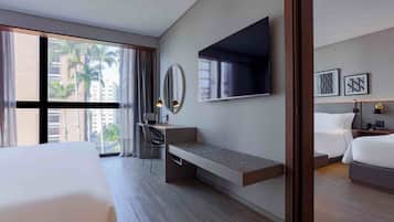 A modern hotel room with a large bed, a desk with a chair, a television, and a view of palm trees through the window.