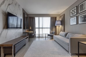 A modern hotel room with a marble wall, a flat-screen TV, a sofa, a dining table, and a lamp.