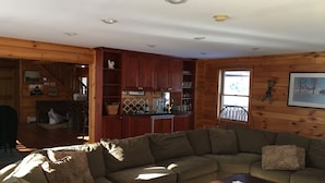 Una sala de estar acogedora con un amplio sofá modular, una mesa de centro de madera y una zona de cocina con armarios de madera y un ventanal.