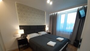 A neatly made bed with pillows and a blanket, a bedside lamp, a nightstand, and a window with curtains. - Apartament plaża 41 bros (Gdansk)