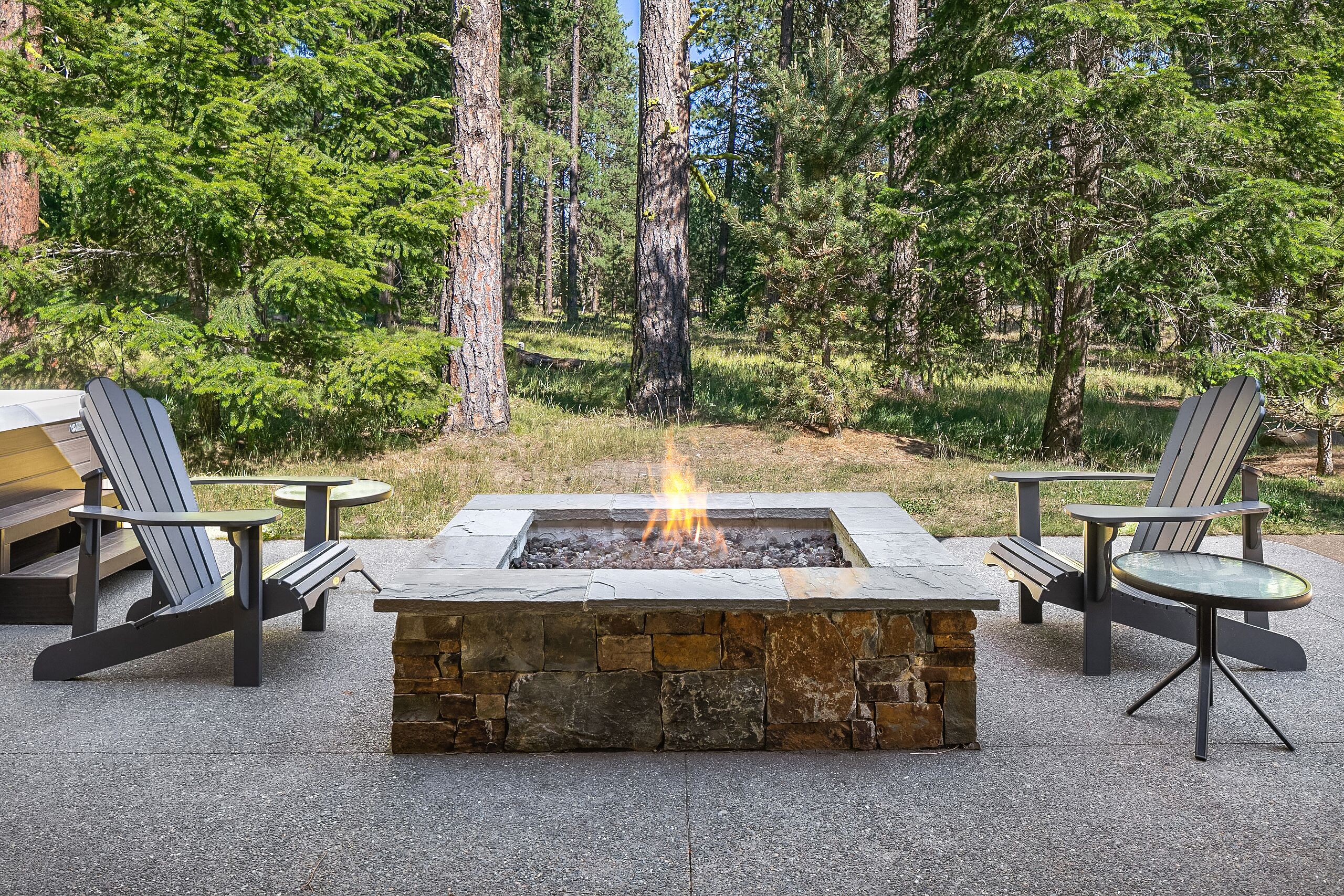 Un foyer en pierre, agrémenté de deux fauteuils Adirondack et d’une table, le tout entouré d’une forêt.