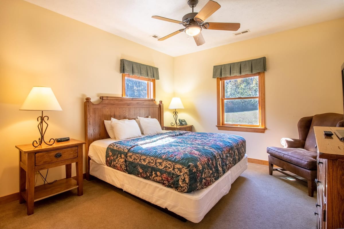 A bedroom with a large bed, two bedside tables with lamps, a ceiling fan, and two windows with curtains.