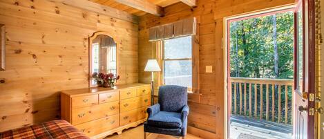 Habitación de cabaña de madera con cama, cómoda, silla y un amplio ventanal con vistas a árboles.