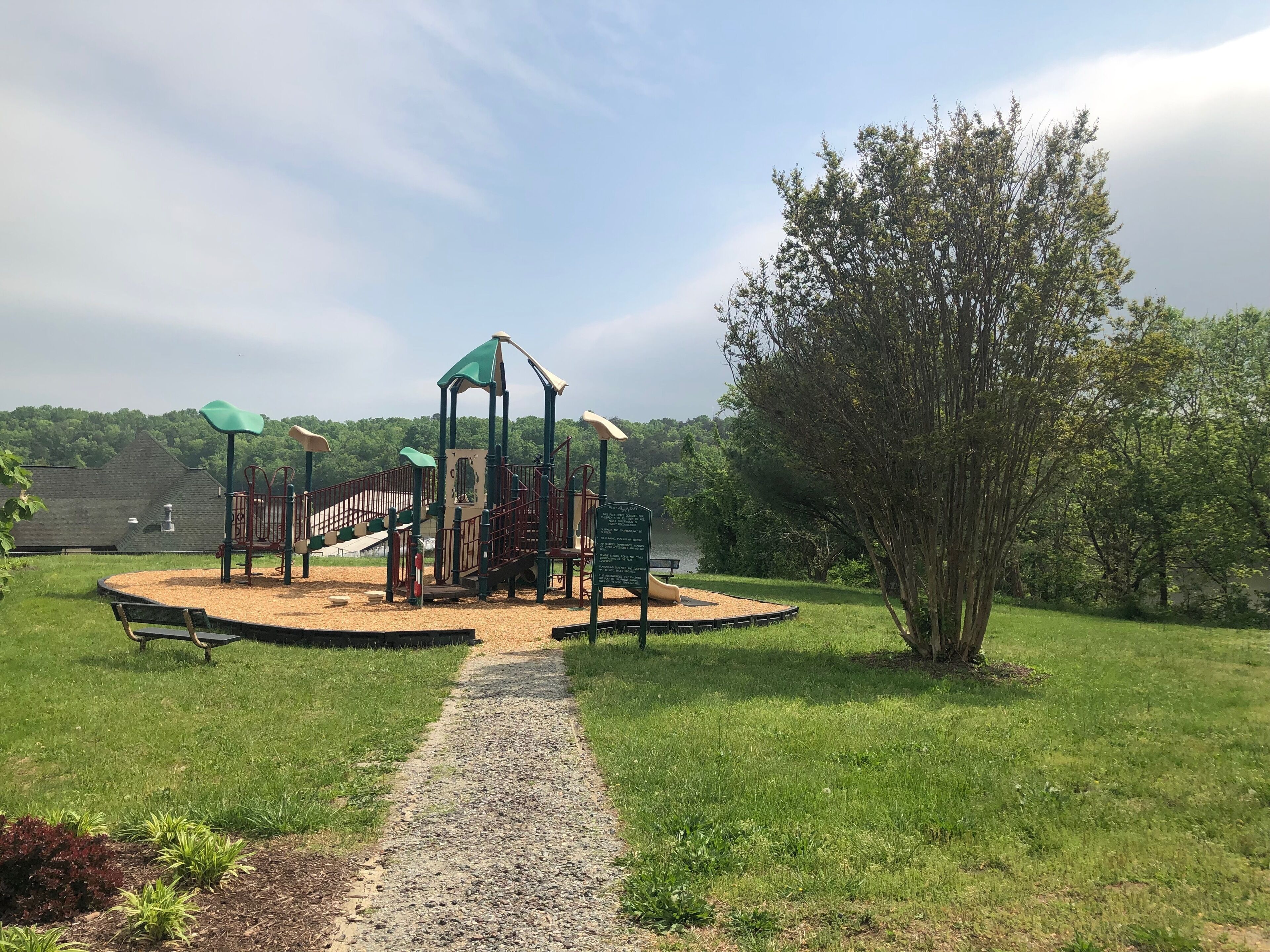 Un parque infantil con un tobogán, estructuras para escalar y una zona cubierta, rodeado de césped y árboles.