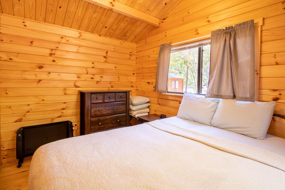 A wooden cabin bedroom with a bed, dresser, and window with curtains.