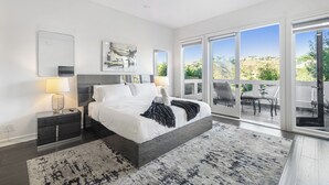 A modern bedroom with a large bed, a nightstand, a bedside lamp, and a view of the outdoors through large sliding glass doors.