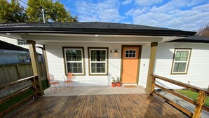 Ein Haus mit einer Holzterrasse, zwei Fenstern, einer Tür und einer kleinen Veranda mit Stühlen und Pflanzen in Töpfen.