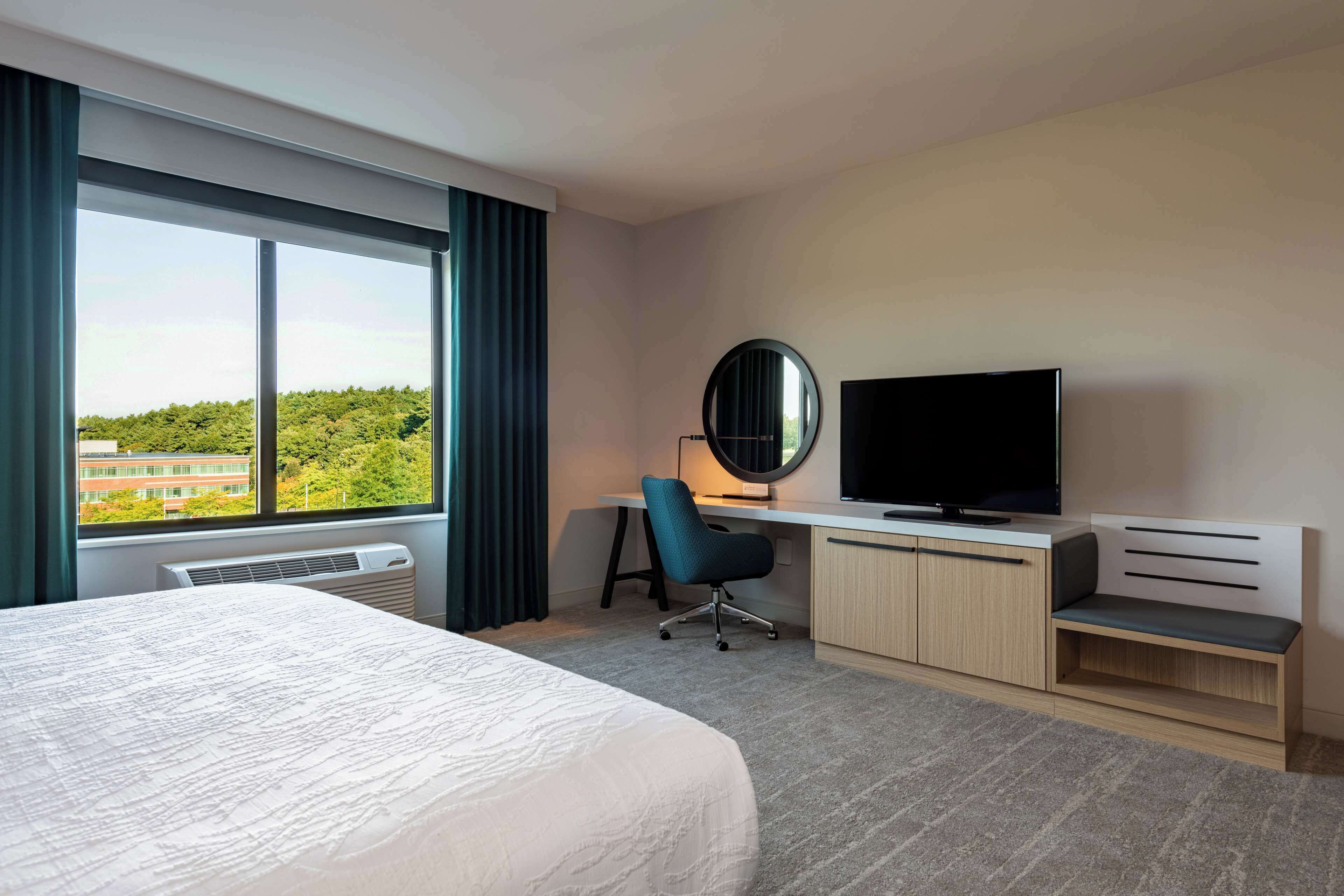 Quarto de hotel com uma cama grande, uma escrivaninha com cadeira, televisão e vista para o verde lá fora.