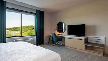Quarto de hotel com uma cama grande, uma escrivaninha com cadeira, televisão e vista para o verde lá fora.