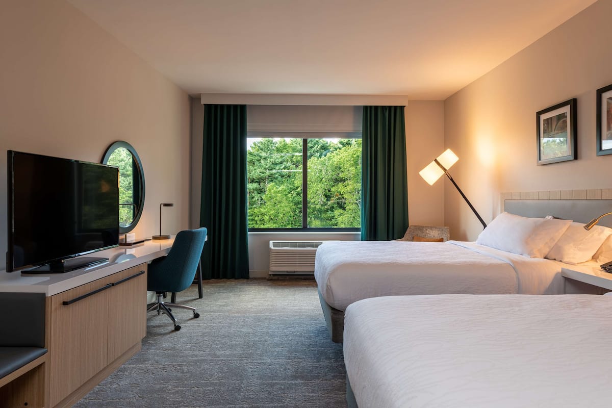 A hotel room with two beds, a desk with a television, a chair, a window with curtains, and a lamp.