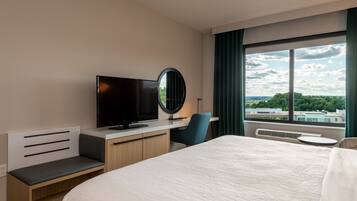 Una habitación de hotel moderna con una cama grande, un escritorio con televisor de pantalla plana, un espejo redondo y vistas de la ciudad a través de amplias ventanas.