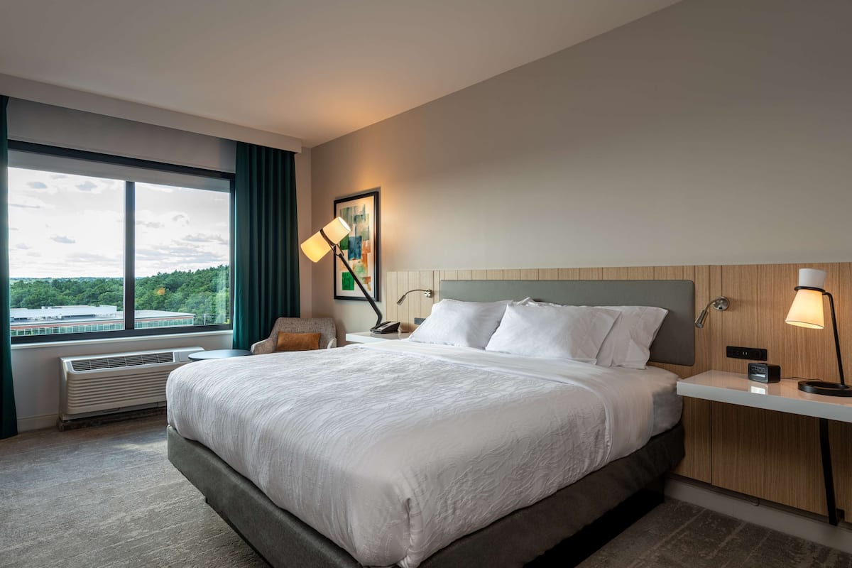 A modern hotel room with a large bed, a desk, a chair, a standing lamp, and a window with a view of greenery and buildings.