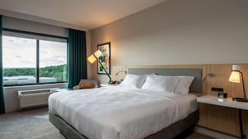 Una camera d'albergo moderna con un letto grande, una scrivania, una sedia, una lampada da terra e una finestra che si affaccia su vegetazione ed edifici.