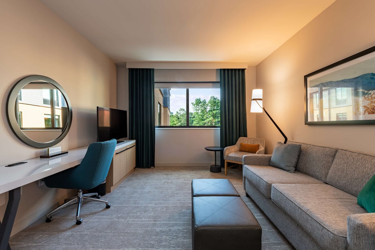 A modern hotel room with a desk, chair, sofa, ottoman, TV, lamp, and a window with curtains.