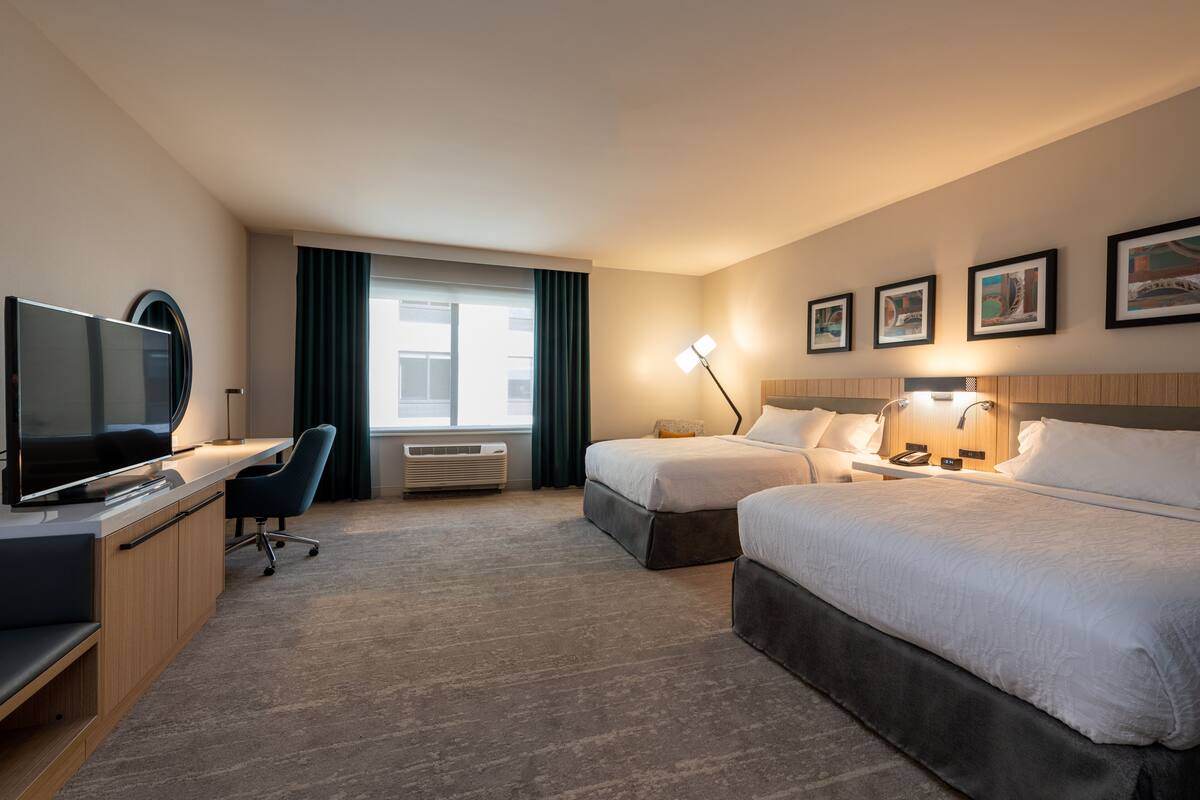 A hotel room with a bed, a desk with a TV, a chair, a window with curtains, and a wall with framed pictures.