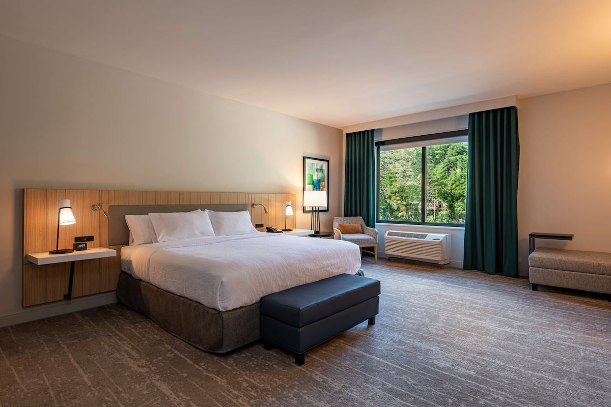 A hotel room with a large bed, bedside tables with lamps, a chair, a bench, and a window with curtains.