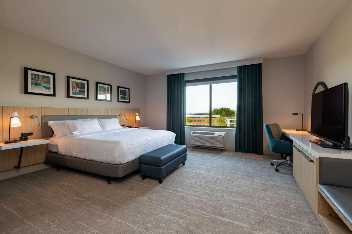 Quarto de hotel com cama, escrivaninha, televisão, cadeira e janela com cortinas.