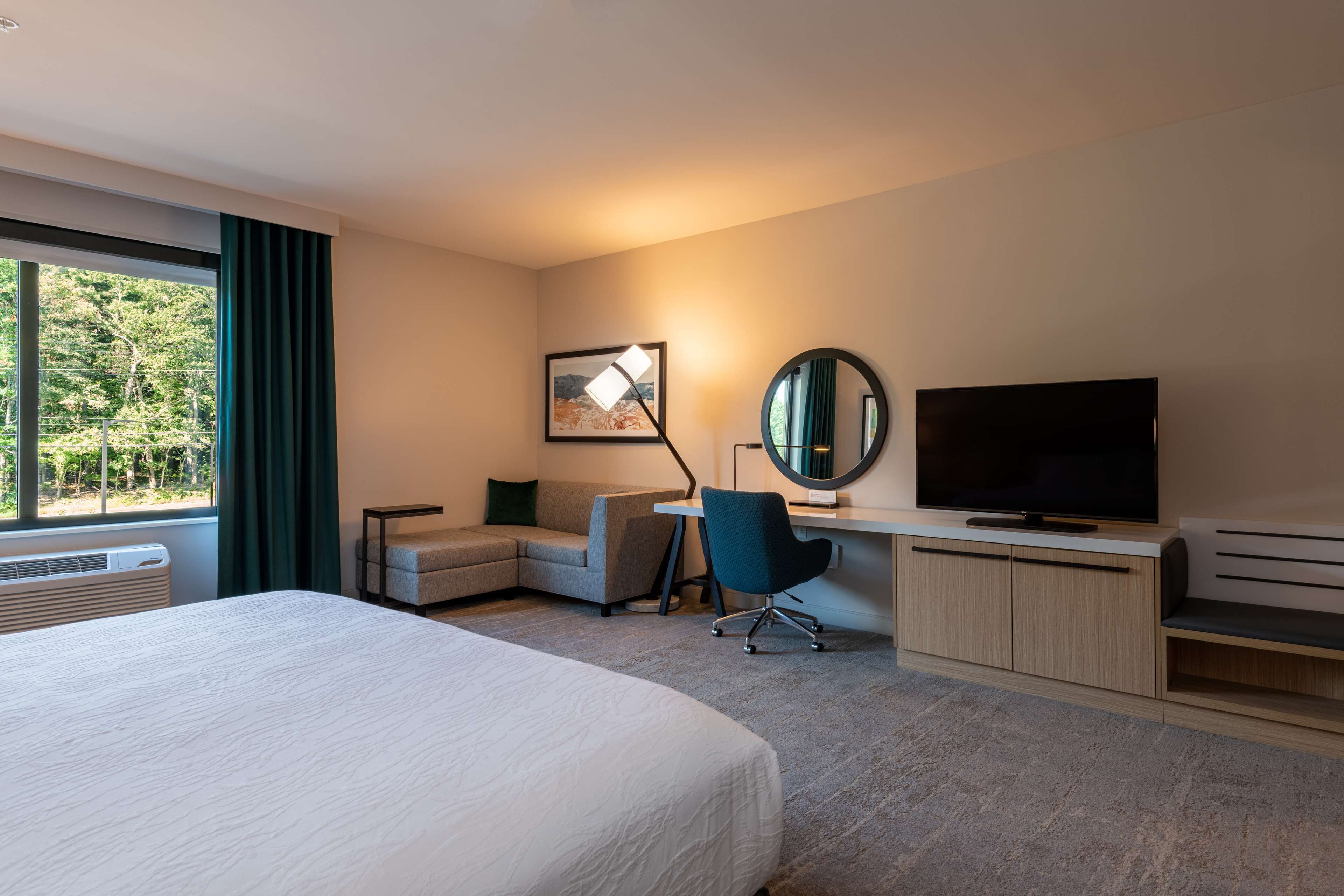 Habitación de hotel con una cama grande, un escritorio con computadora, un televisor y una ventana con cortinas.
