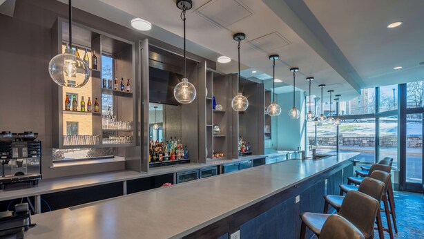 A modern bar area with a long counter, multiple hanging pendant lights, and a variety of bottles on shelves. - Scholar State College, Tapestry Collection by Hilton (State College)