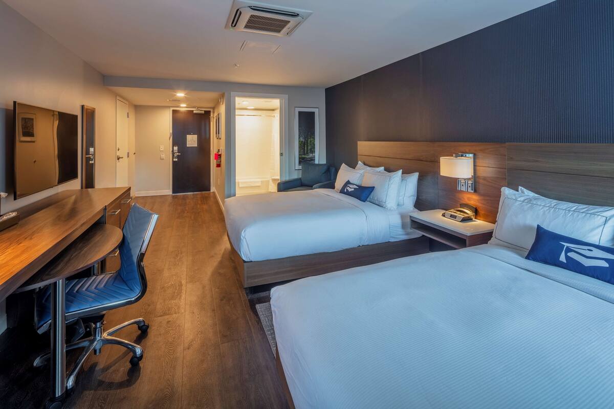 A hotel room with two beds, a desk, and a chair. There is a door leading to a bathroom and a hallway.