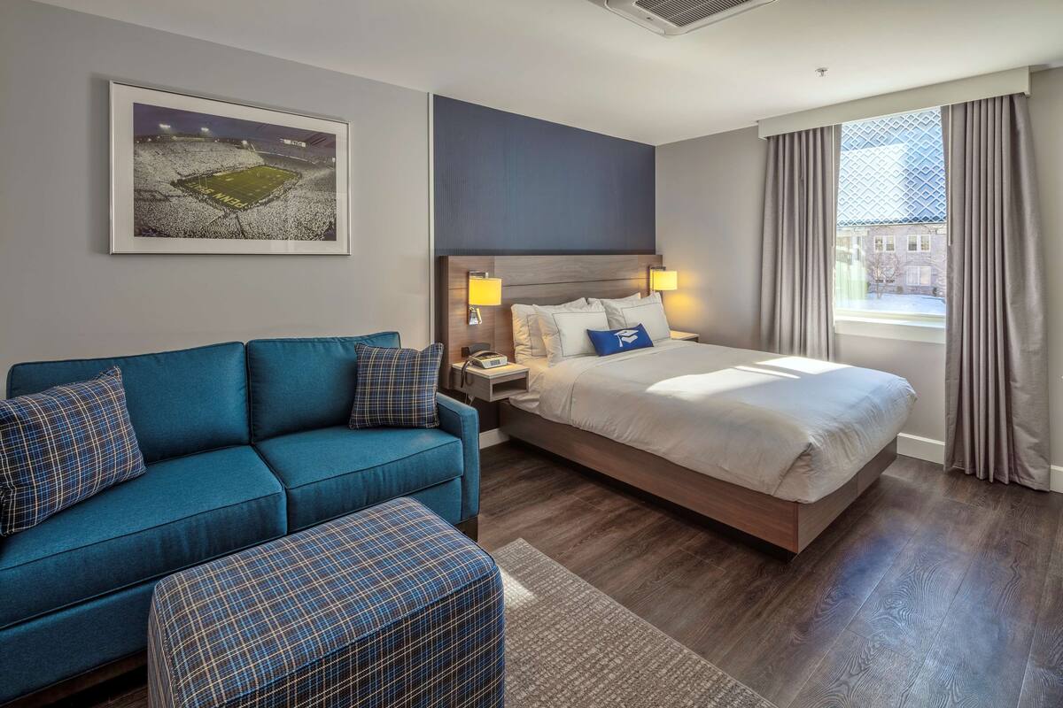A hotel room with a bed, a blue sofa, a matching ottoman, a nightstand, and a framed picture on the wall.