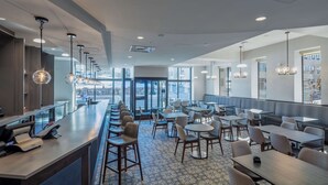 A modern restaurant with a long counter, multiple seating areas, and large windows providing views of the outside. - Scholar State College, Tapestry Collection by Hilton (State College)