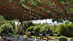 En uteservering med en pergola, bord som är dukade med tallrikar, glas och bestick, omgiven av grönska.