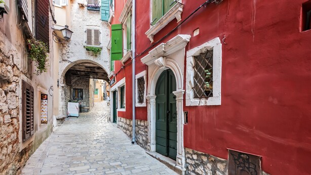 A narrow cobblestone street flanked by buildings with stone facades and green shutters.