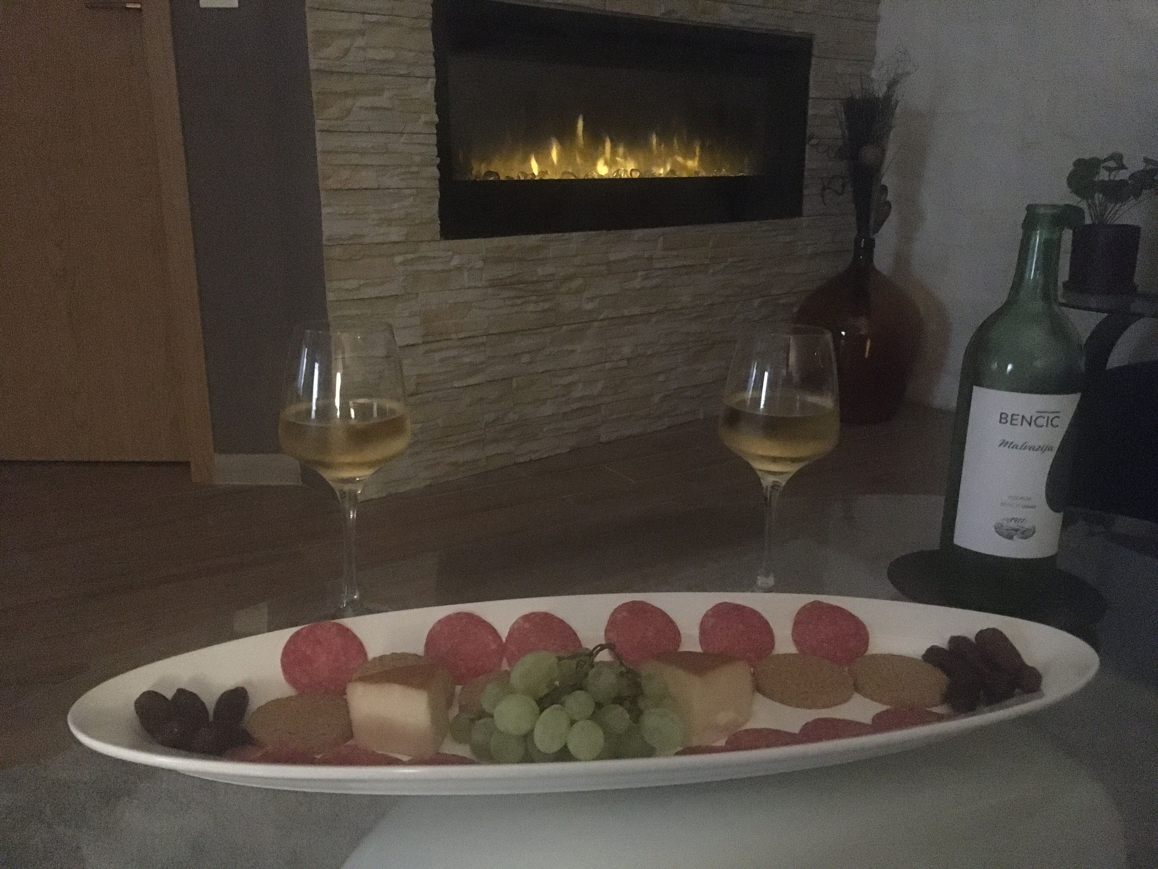 Une table à manger avec une assiette blanche remplie d’un assortiment de fruits et deux verres de vin blanc, une bouteille de vin Bencic, et une cheminée en arrière-plan.