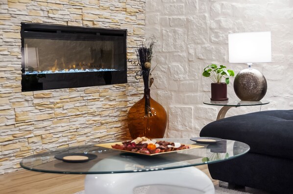 A modern living room with a stone wall, a fireplace, a glass coffee table with snacks, a dark sofa, and a potted plant. - Luxury Apartment Kastela Antika (Rovinj)
