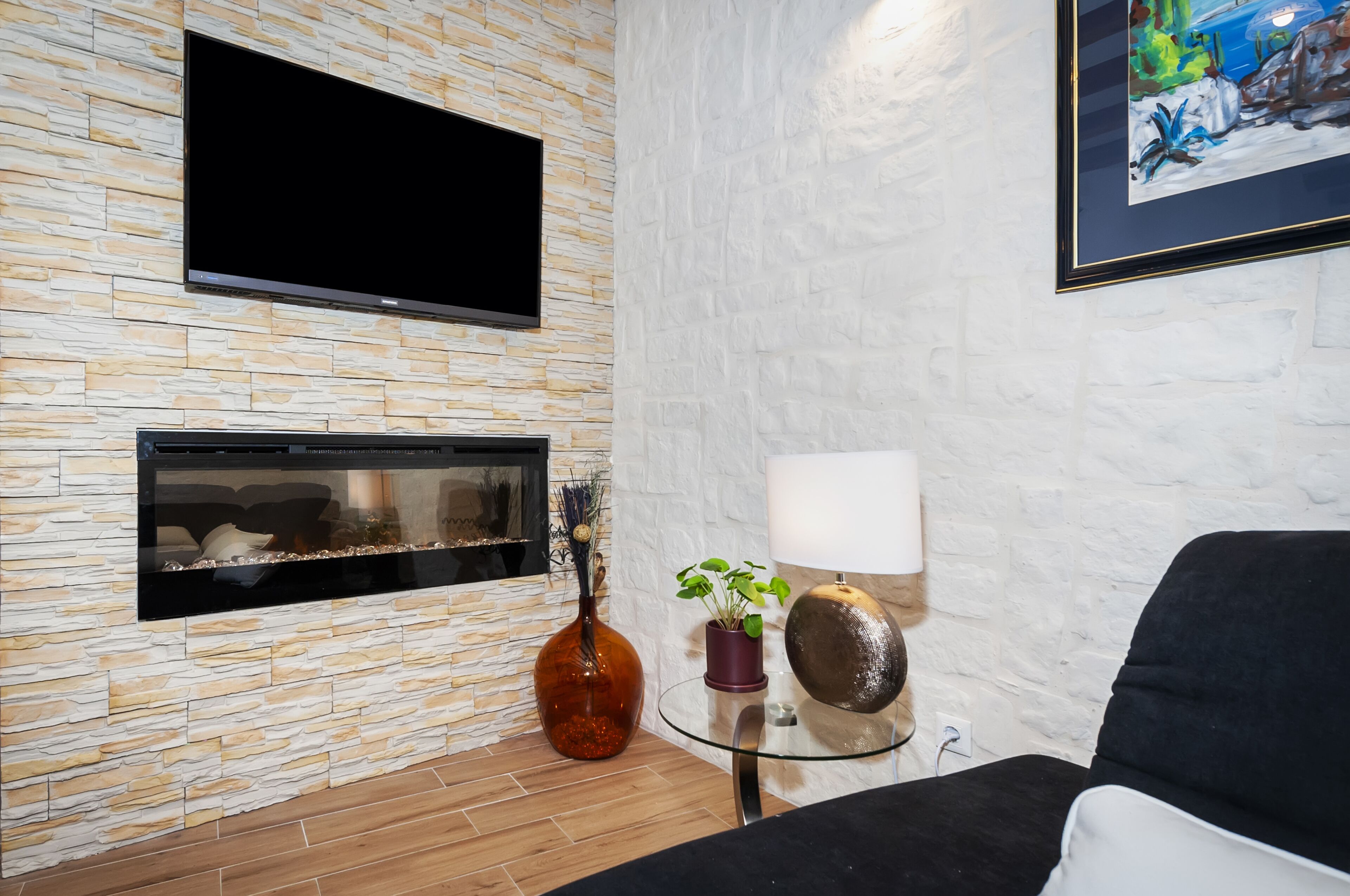 A modern living room with a stone-textured wall, a flat-screen TV, a fireplace, a glass coffee table, a potted plant, a lamp, and a black sofa.