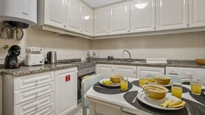 A modern kitchen with white cabinets, a built-in oven, a coffee maker, and a dining area with a table set for two. - Netos Apartment - By WeHostMadeira (Funchal)