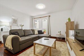 Ein modernes Wohnzimmer mit einer grauen Eckcouch, einem weißen Couchtisch, einer TV-Konsole mit einem Flachbildfernseher und einem kleinen Beistelltisch mit einer Pflanze.